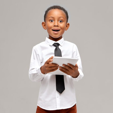 Astonished dark-skinned little boy wearing white shirt and black tie enjoying high speed wireless internet connection on digital tablet having surprised amazed look, watching cartoon online
